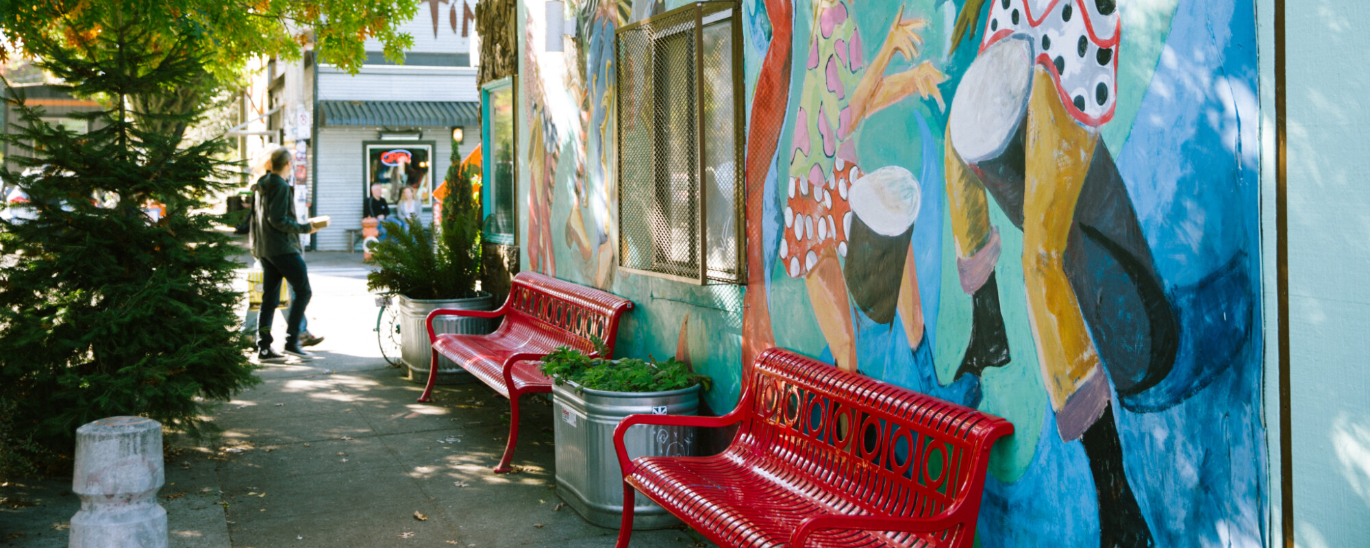A sidewalk scene featuring trees (left) and the exterior wall of a building that has a vibrant mural featuring people of diverse ages and cultures dancing (right).