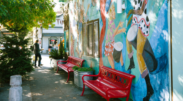 Una escena en la acera con árboles (izquierda) y la pared exterior de un edificio que tiene un vibrante mural con personas de diversas edades y culturas bailando (derecha).