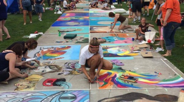 Spectators watch artists create chalk art on a sidewalk path.