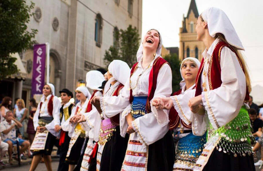 Women wearing traditional Greek clothing joyously dance on a downtown street.