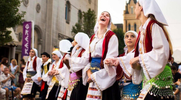 Women wearing traditional Greek clothing joyously dance on a downtown street.