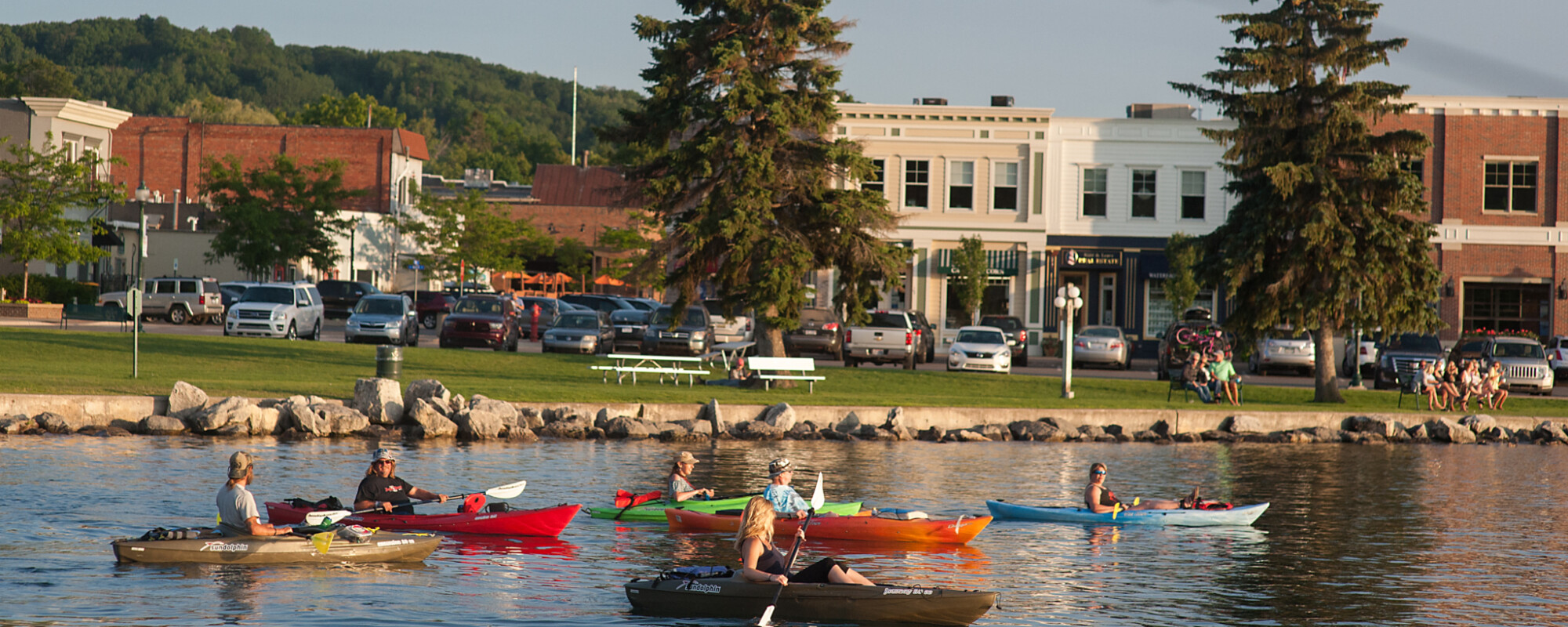 People in kayaks on a lake with historic retail storefronts in the background.