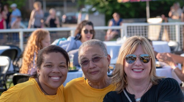 Three women smile while attending an outdoor community event.