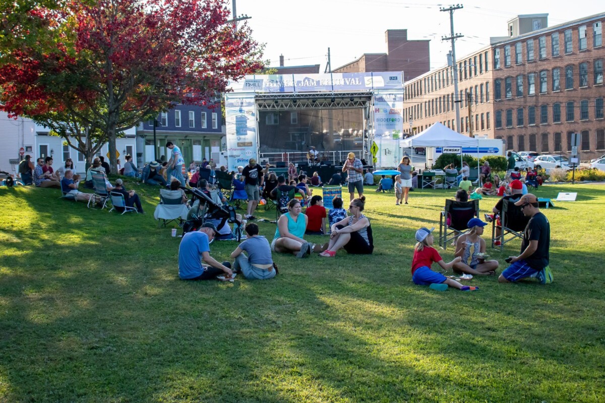 People gather in small groups on a shady grass lawn in front of a concert stage.