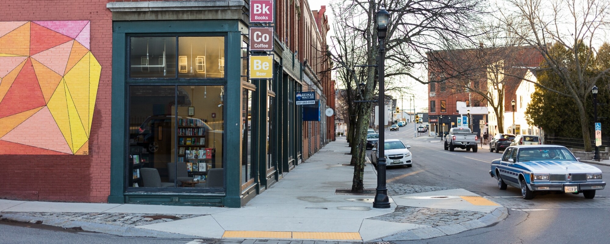 View of a downtown street corner. From left to right: two-story, historic brick buildings; a wide, tree-lined sidewalk with curb ramps at intersections; and a two-lane roadway with vehicles traveling.