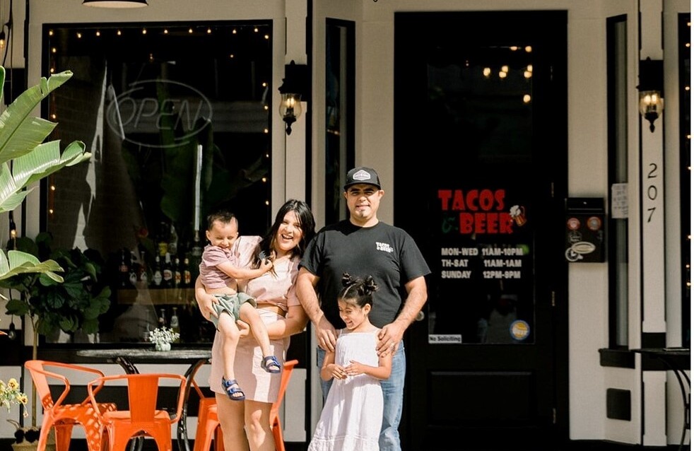 A family of two adults and two young children pose in front of their business, Tacos & Beer.