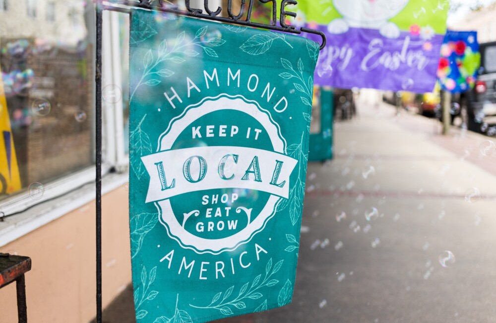 A vibrant teal banner hangs from a black metal stand placed on a sidewalk. The banner reads: "Hammond America: Keep it Local. Shop, Eat, Grow"