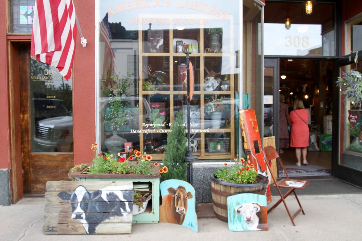 A retail storefront display featuring farm-themed items thoughtfully positioned in the large glass windows and on the sidewalk. Text on the window reads: America's Largest Craftsman Co-Op.