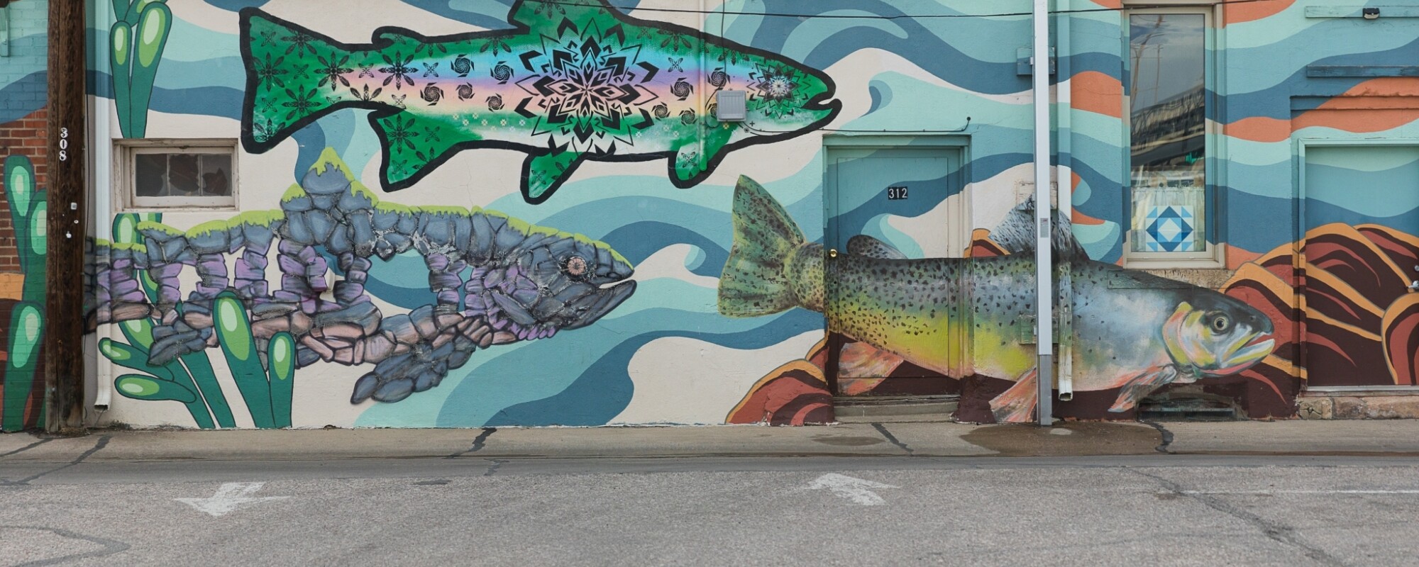 A mural featuring larger than life trout painted in vibrant colors and swirls of blue representing water cover the exterior of a commercial building.