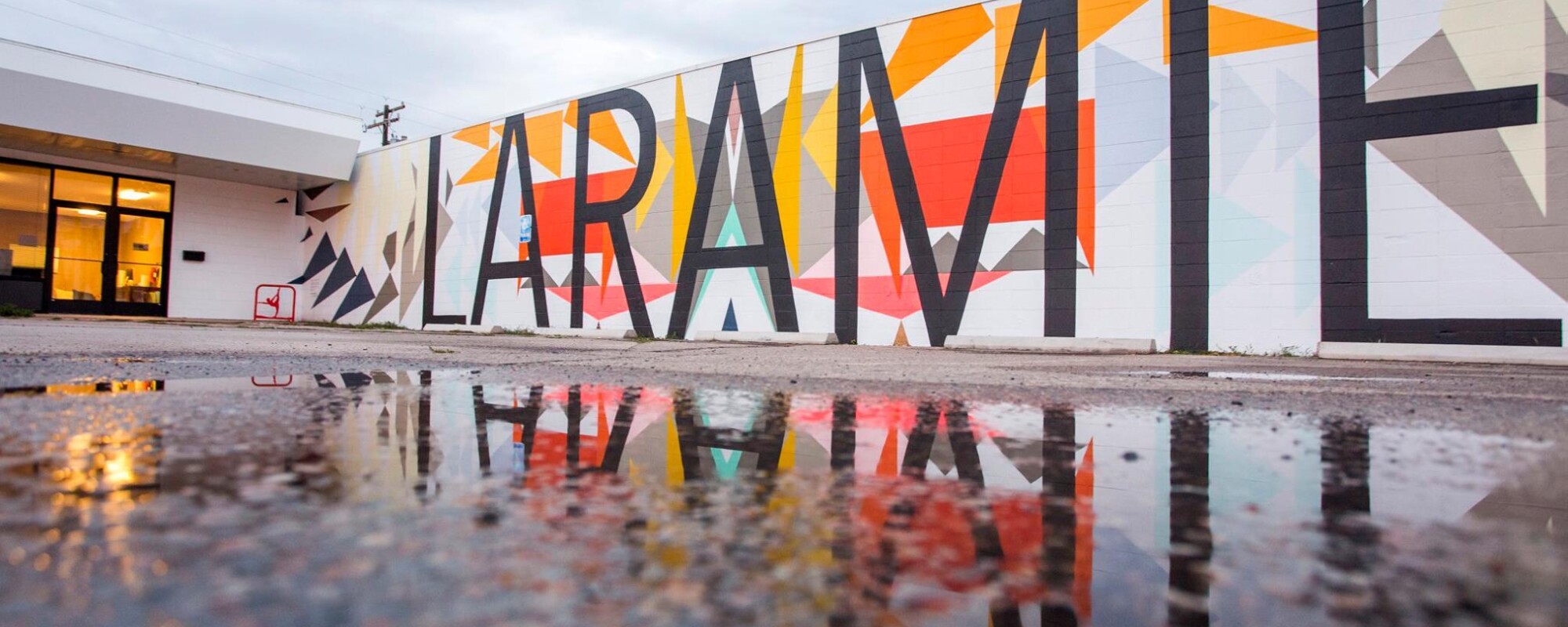 Mural featuring the word "LARAMIE" is reflected in the puddle of a parking lot.