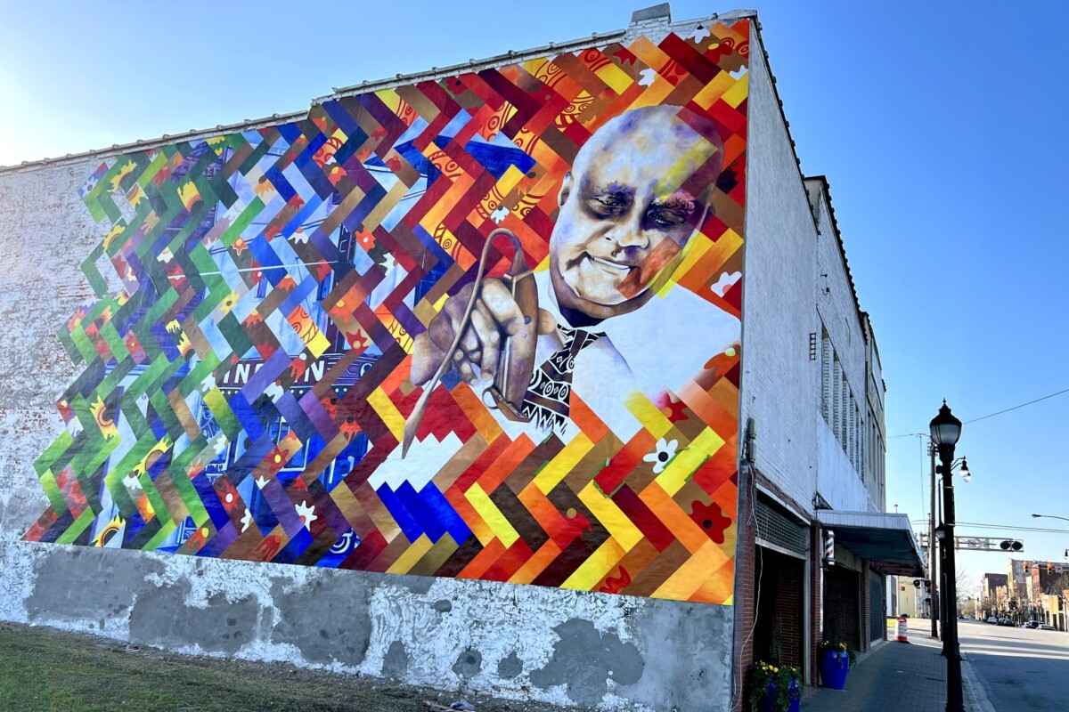 A colorful mural featuring the depiction of an African American barber cutting hair installed on the side of a historic brick building.