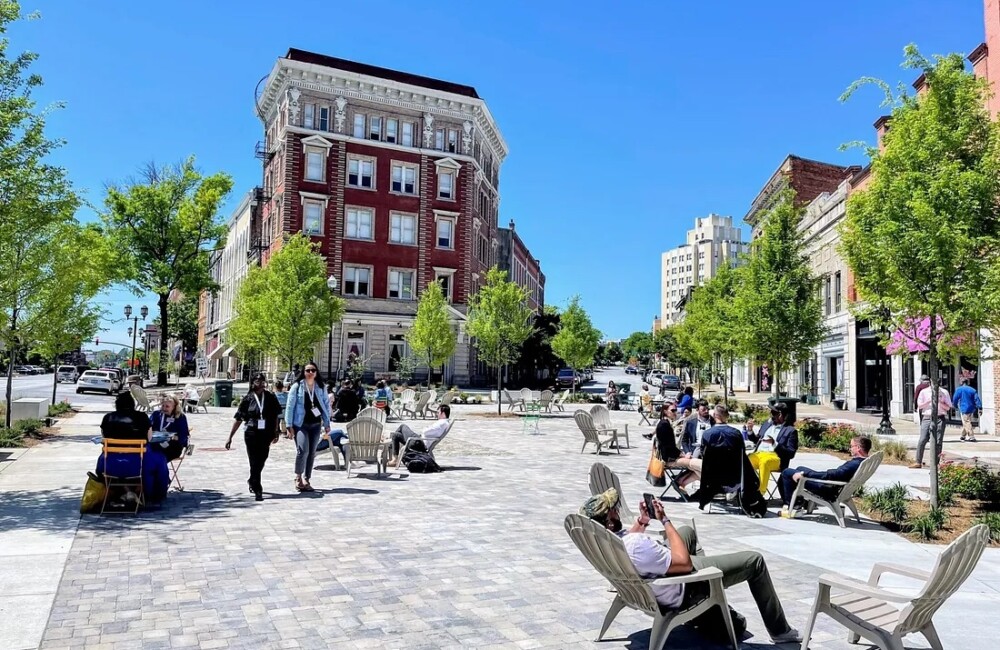 People lounge in chairs, sit at bistro tables, and stroll through a large plaza dotted with trees and surrounded by historic brick buildings.