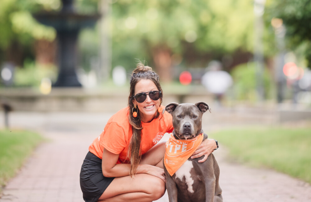 A woman wearing a bright orange shirt crouches next to her dog while in a park.