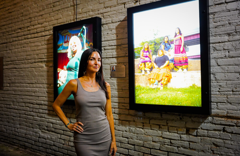 A woman, with her hand placed on her hip, looks confidently ahead while standing in front of illuminated light boxes featuring portraits of citizens of the Muscogee (Creek) Nation.