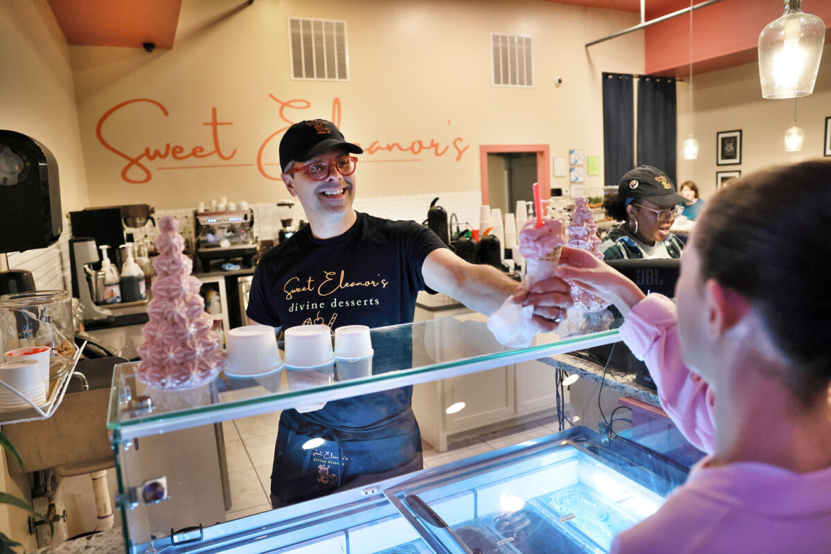 Inside an ice cream shop, a man smiles while handing an ice cream cone to a customer.