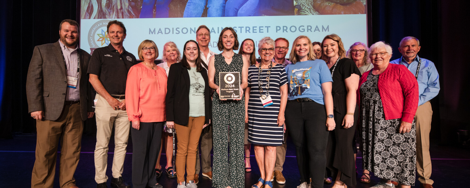 A large group of people stand on stage; the person in the center holds a plaque.