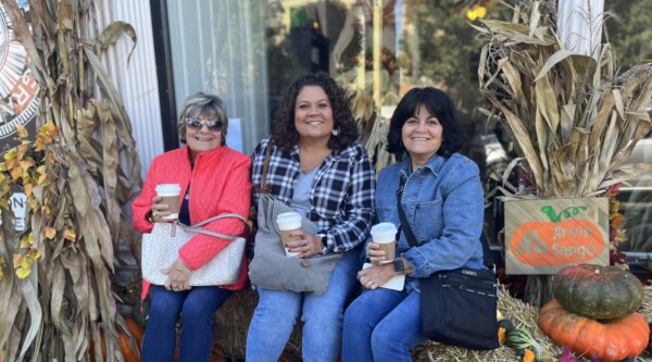 Three women hold cups of coffee while sitting on a hay bale.