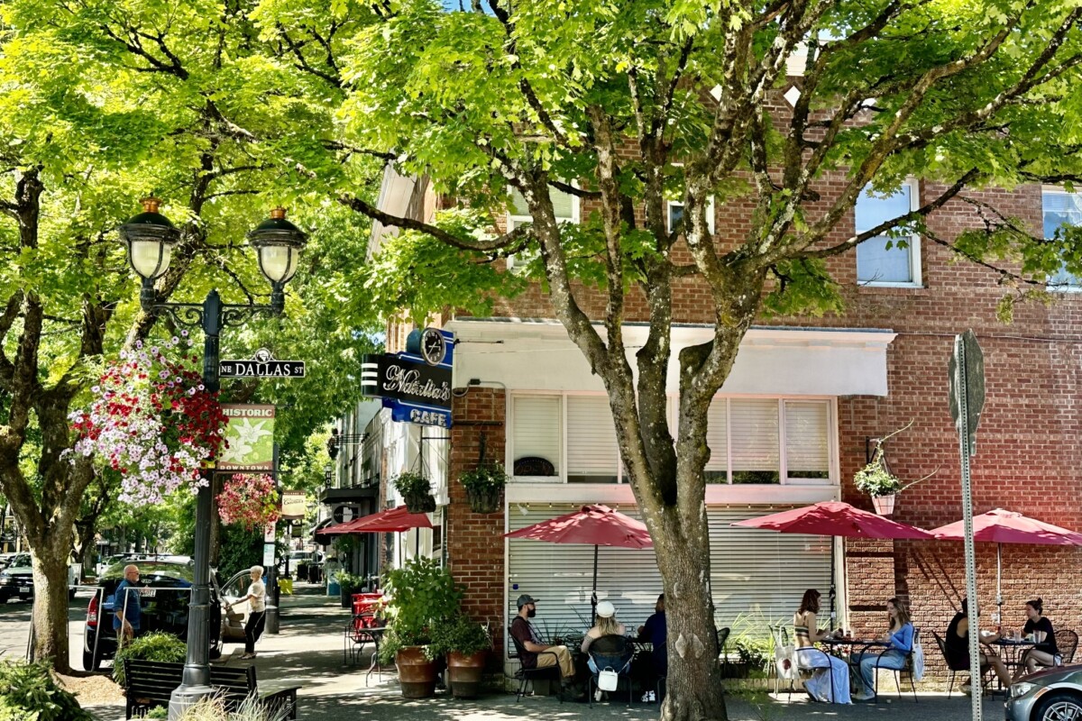 On a sunny day, diners enjoy outdoor sidewalk seating under the shade of trees and meals from a café located in a historic brick building.