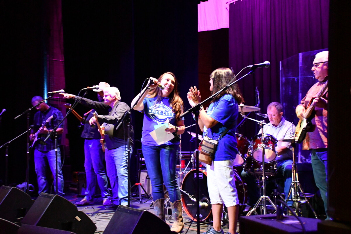 A woman stands center stage while speaking into a microphone and surrounded by a band.