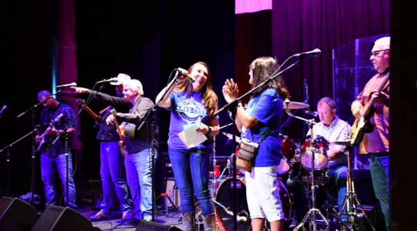A woman stands center stage while speaking into a microphone and surrounded by a band.