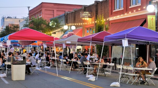 People gather around tables set-up underneath brightly colored pop-up canopy tents that have been arranged in a single line down the middle of a downtown street.