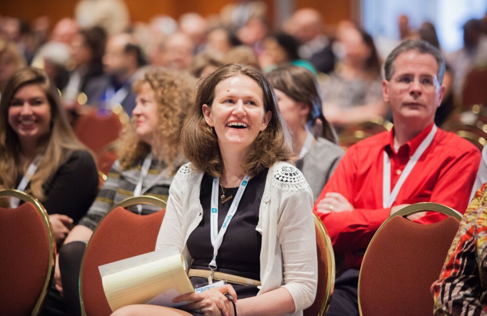 A woman smiles while sitting in the audience of a speaking event.