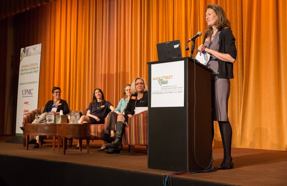 A woman makes remarks from behind a podium while, to her right, a panel of speakers sit in chairs.