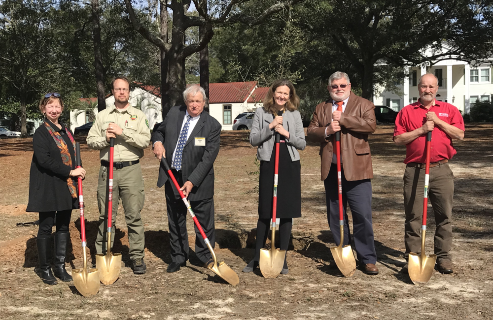 Six people stand shoulder to shoulder in the clearing of a park holding shovels.