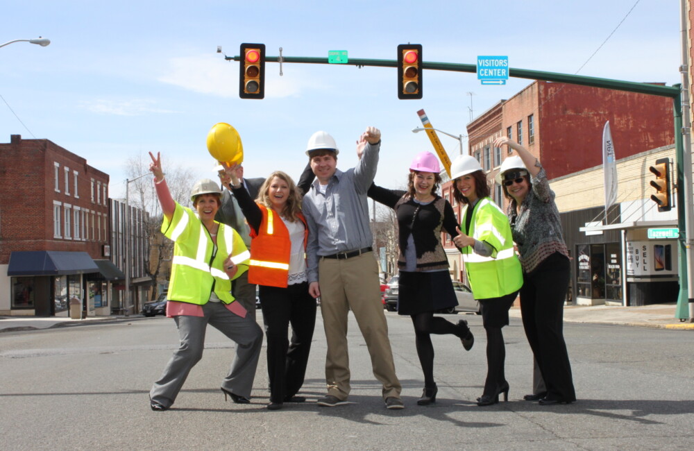 A small group of people wearing hi-vis vests and hard hats stand in the middle of an intersection celebrating the kick-off of streetscape improvement project.