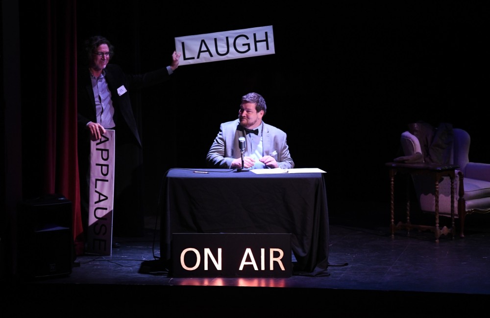On a theater state, a man sits behind a table that is draped in black cloth. In front of the table, an illuminated sign reads "ON AIR." Stage right, another man holds up a sign reading "LAUGH" with his left hand; in his right hand, another sign reading "APPLAUSE" points downward.