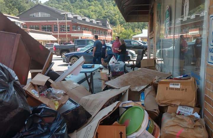 A large pile of flood damaged boxes, full trash bags, and miscellaneous material covers a sidewalk. People and buildings are visible in the background.