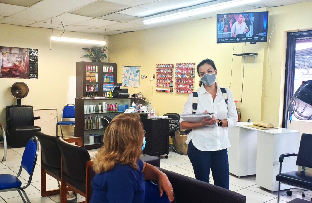 Inside a beauty salon, a woman holds a clipboard while speaking with a woman who is seated.