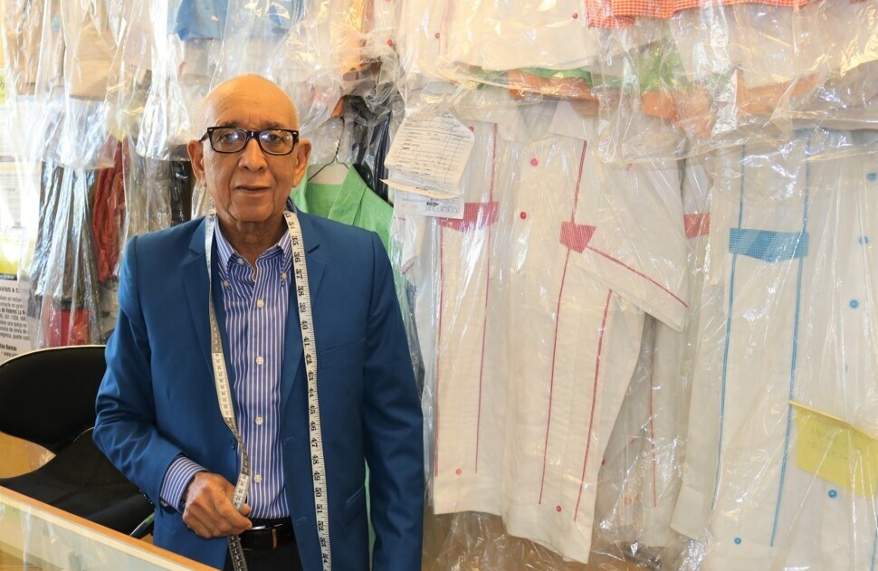 A man wearing a blue suit with a fabric measuring tape draped around his shoulders stands in front of a wall of clothes encased in plastic garment bags.