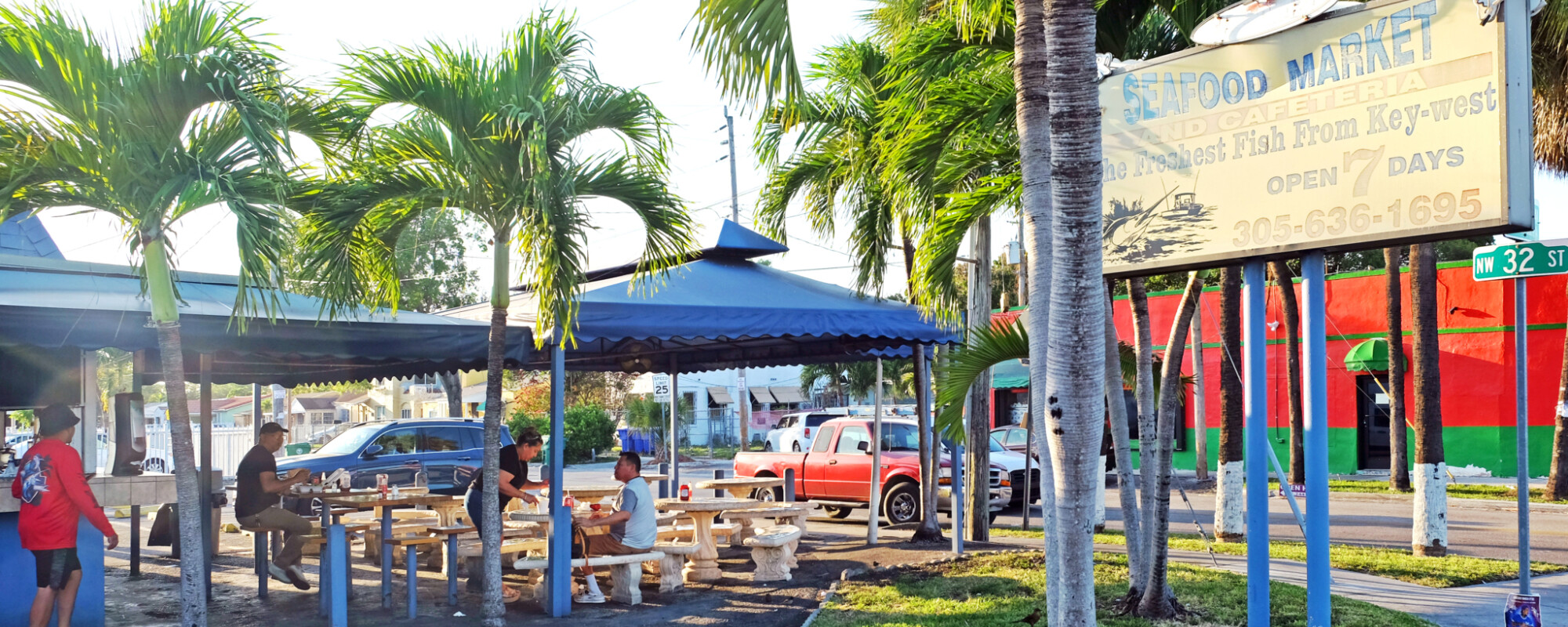 People sit in an outdoor dining area with picnic tables, canopies, and palm trees.