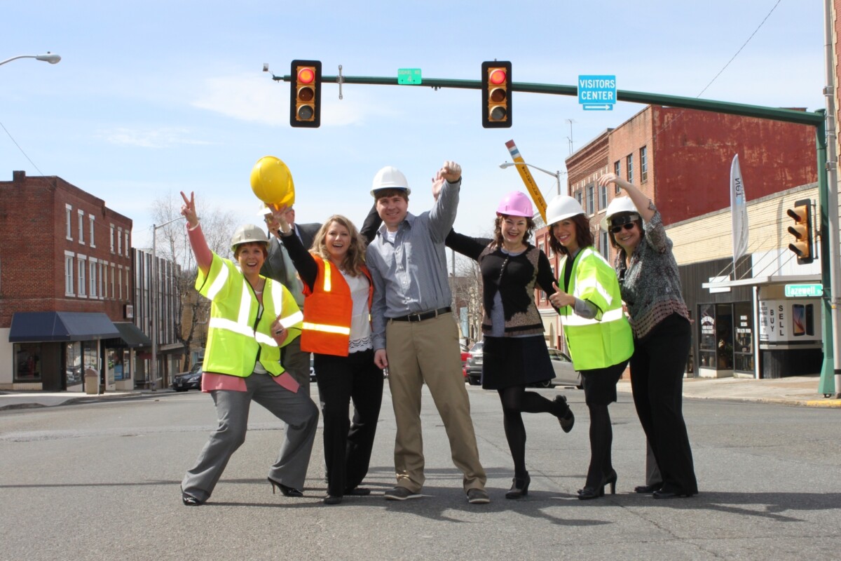 A small group of people wearing hi-vis vests and hard hats stand in the middle of an intersection celebrating the kick-off of streetscape improvement project.