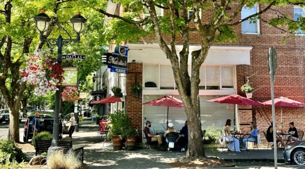 En un día soleado, los comensales disfrutan de los asientos al aire libre bajo la sombra de los árboles y de las comidas de una cafetería situada en un histórico edificio de ladrillo.