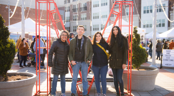 Four people stand shoulder to shoulder in front of a large red "M" structure. Behind them, a plaza is filled with visitors and white pop-up canopy tents staffed by vendors participating in a spring bazaar event.