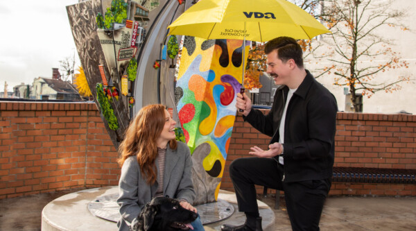 A man holds a bright yellow umbrella while chatting with a woman near a colorful sculpture.