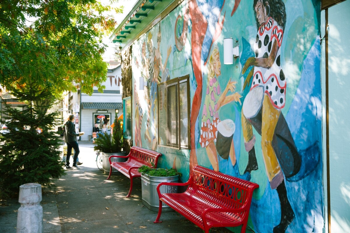 Una escena en la acera con árboles (izquierda) y la pared exterior de un edificio que tiene un vibrante mural con personas de diversas edades y culturas bailando (derecha).