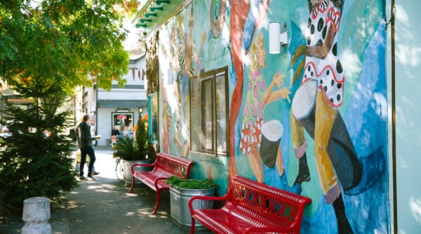 Una escena en la acera con árboles (izquierda) y la pared exterior de un edificio que tiene un vibrante mural con personas de diversas edades y culturas bailando (derecha).