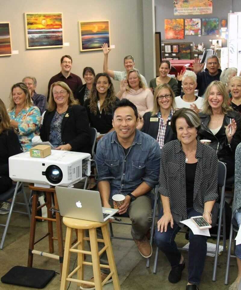 Large group of people gathered in an office space smiling at the camera in front of a projector.