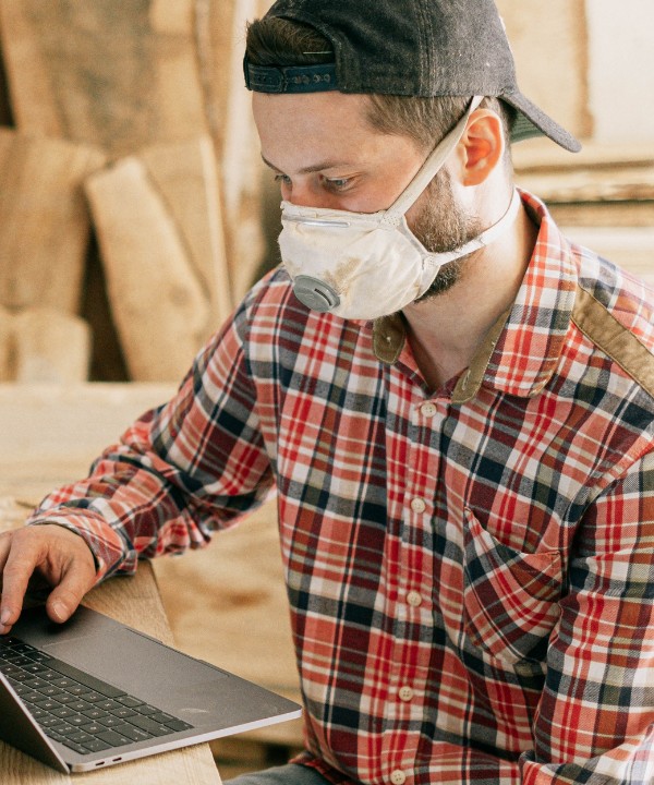 Man wearing backward cap, mask, and flannel shirt sits at a desk in front of a computer.