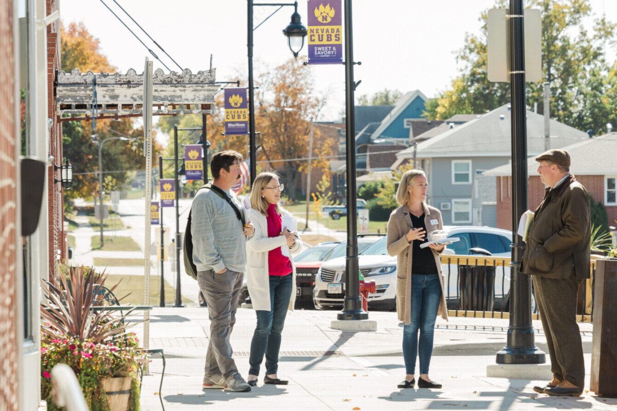 Four people engage in conversation while standing in a downtown area; in the background a theater marquee, houses, and street banners are visible.