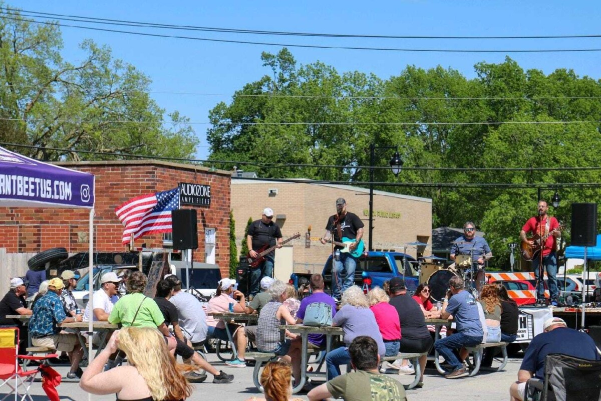 People sit at tables set-up on a street and chat while listening to a band performing on a stage.