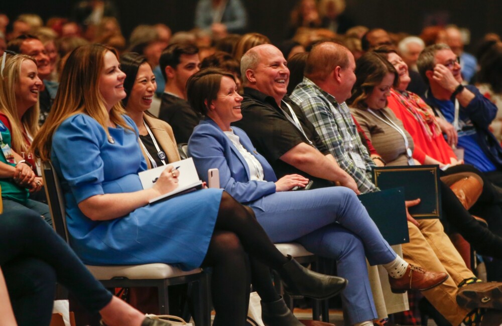 Gente sentada al estilo de un teatro en una gran sala, riendo junto a un orador (que no aparece en la foto).