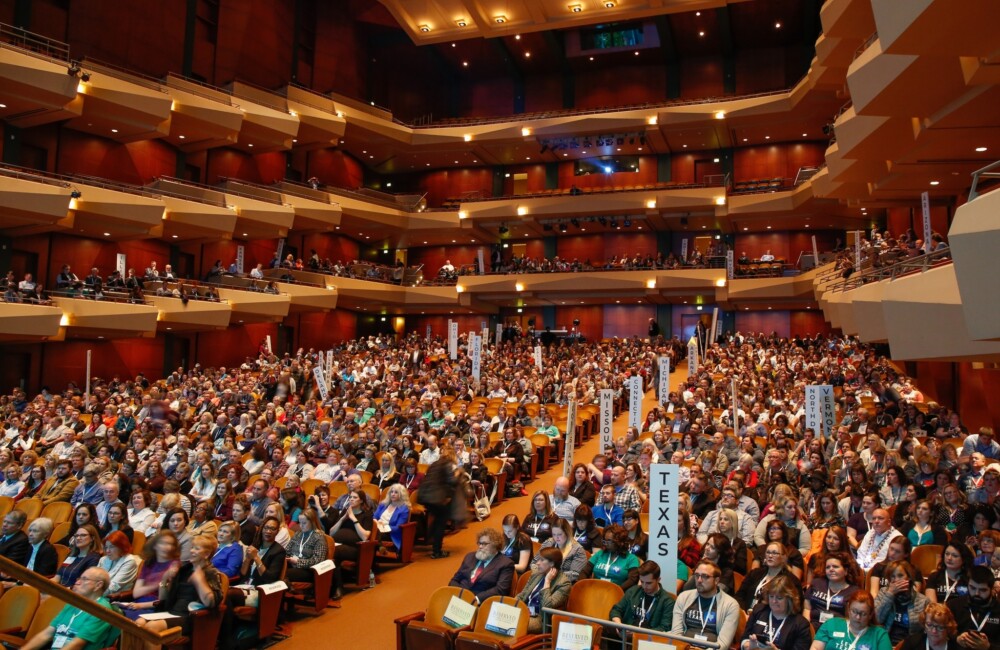 Más de 1.000 personas se reúnen en un gran auditorio con butacas de teatro y de palco; algunas sostienen carteles con los nombres de los estados de Estados Unidos.