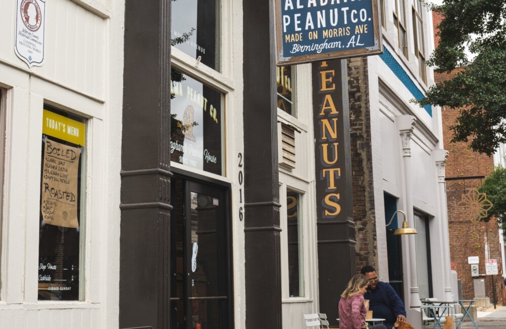 Two people sit at a table on the sidewalk outside of a retail store housed within a historic brick building. The sign hanging from the exterior of the building reads "Alabama Peanut Co."