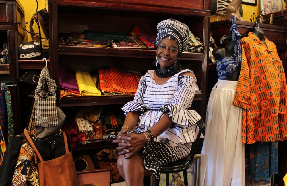 A Black woman sits in a chair and smiles while surrounded by the colorful African clothing on display in her retail store.