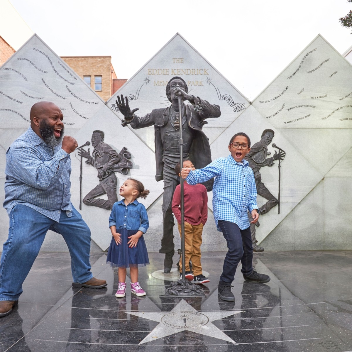 A father and his three young children sing and dance while visiting the Eddie Kendrick Memorial; bronze statues of the singer are visible in the background.