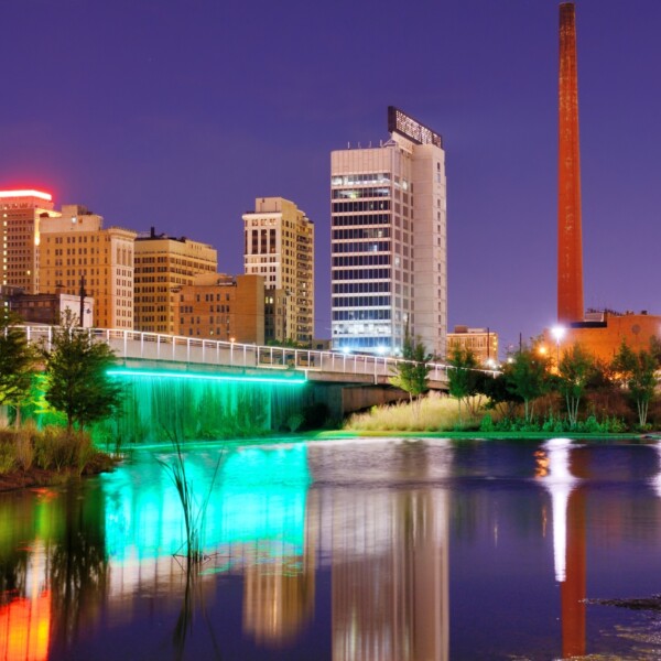 The underside of a bridge is illuminated with teal lights; the city skyline and rust-colored smoke stacks that rise in the background are reflected in the river that flows gently in the foreground.